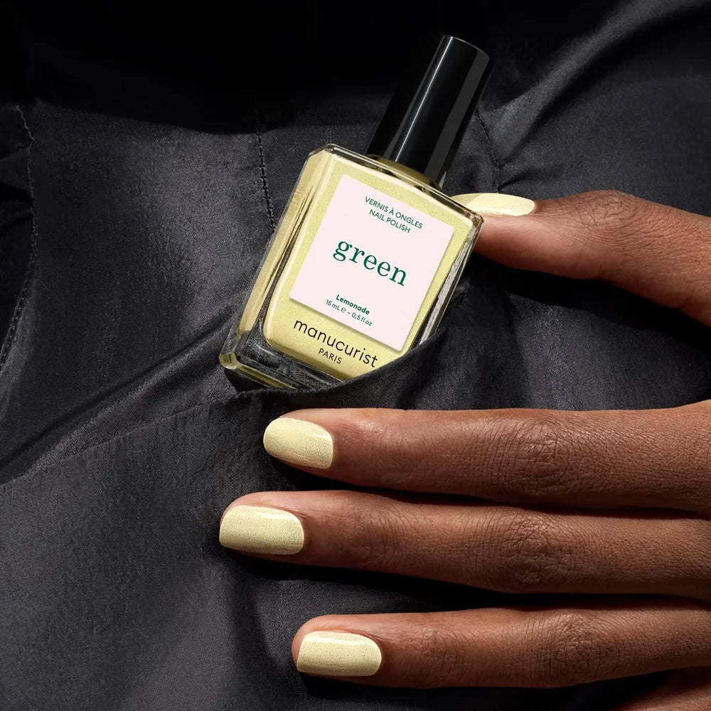 Hand with pastel yellow manicured nails holding Manucurist Paris Green nail polish in Lemonade shade against luxurious black silk fabric