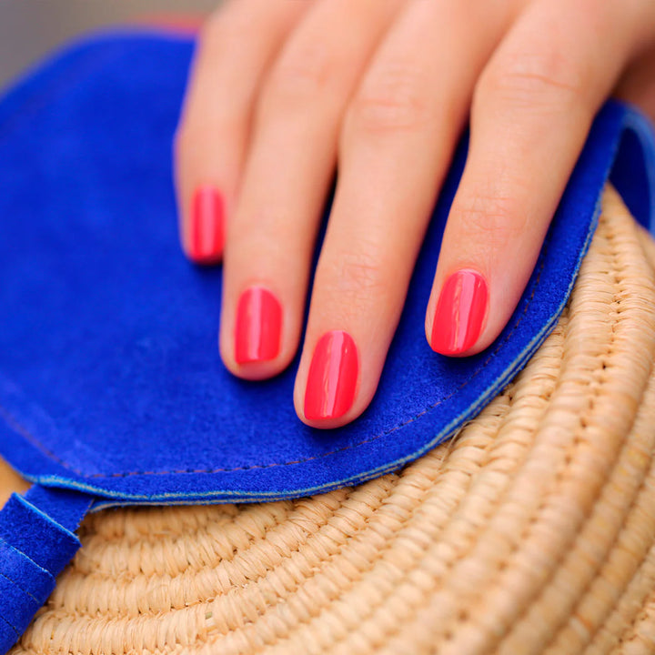Hand mit korallrosa manikürten Nägeln ruht auf blauer Veloursoberfläche und zeigt das Finish des Manucurist Bougainvillea Nagellacks